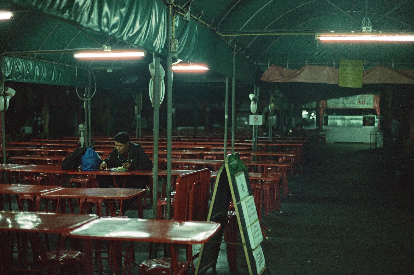 A man sits alone at a red table in an empty outdoor restaurant at night.
