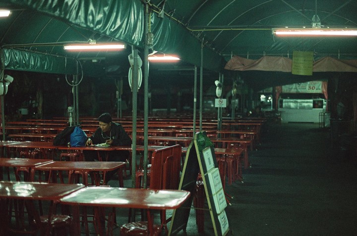 A man sits alone at a red table in an empty outdoor restaurant at night.