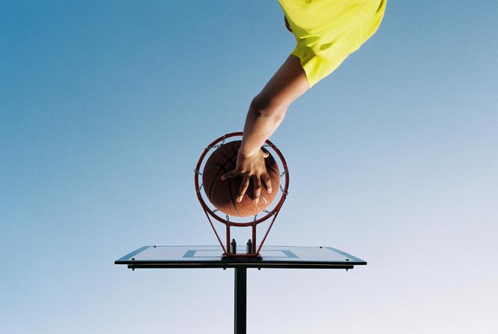A person's arm and a basketball are visible above a basketball hoop against a clear blue sky.