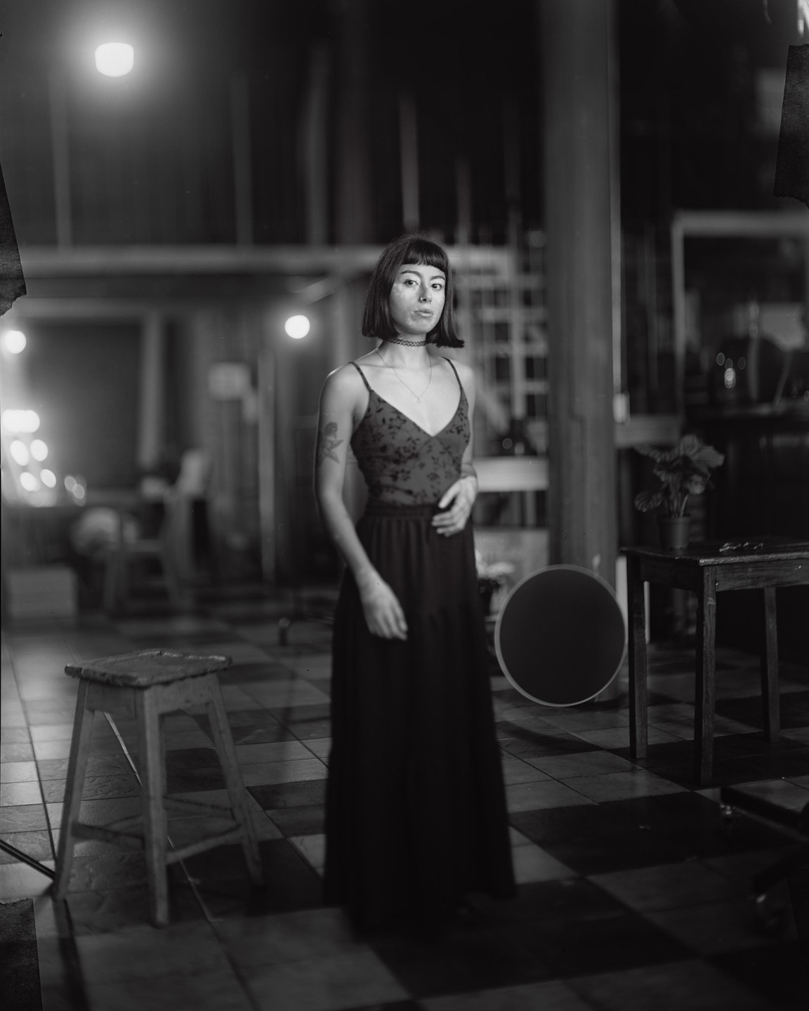 A woman with short hair and a dark dress stands in a dimly lit room with a checkered floor.