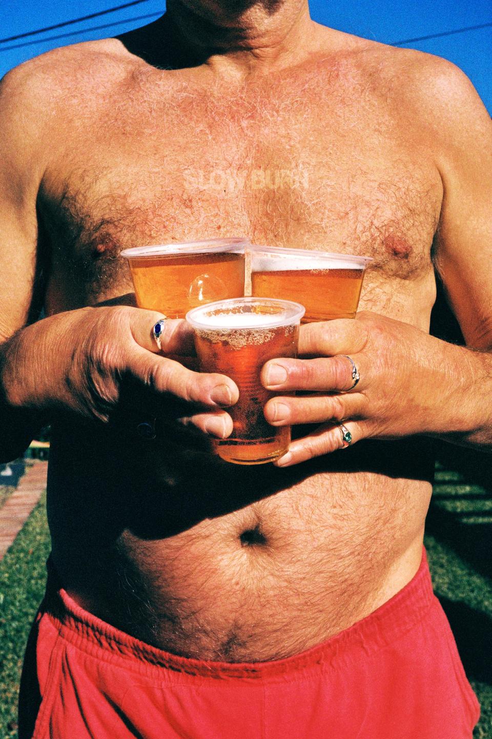 A shirtless man in red shorts holds three plastic cups of beer.