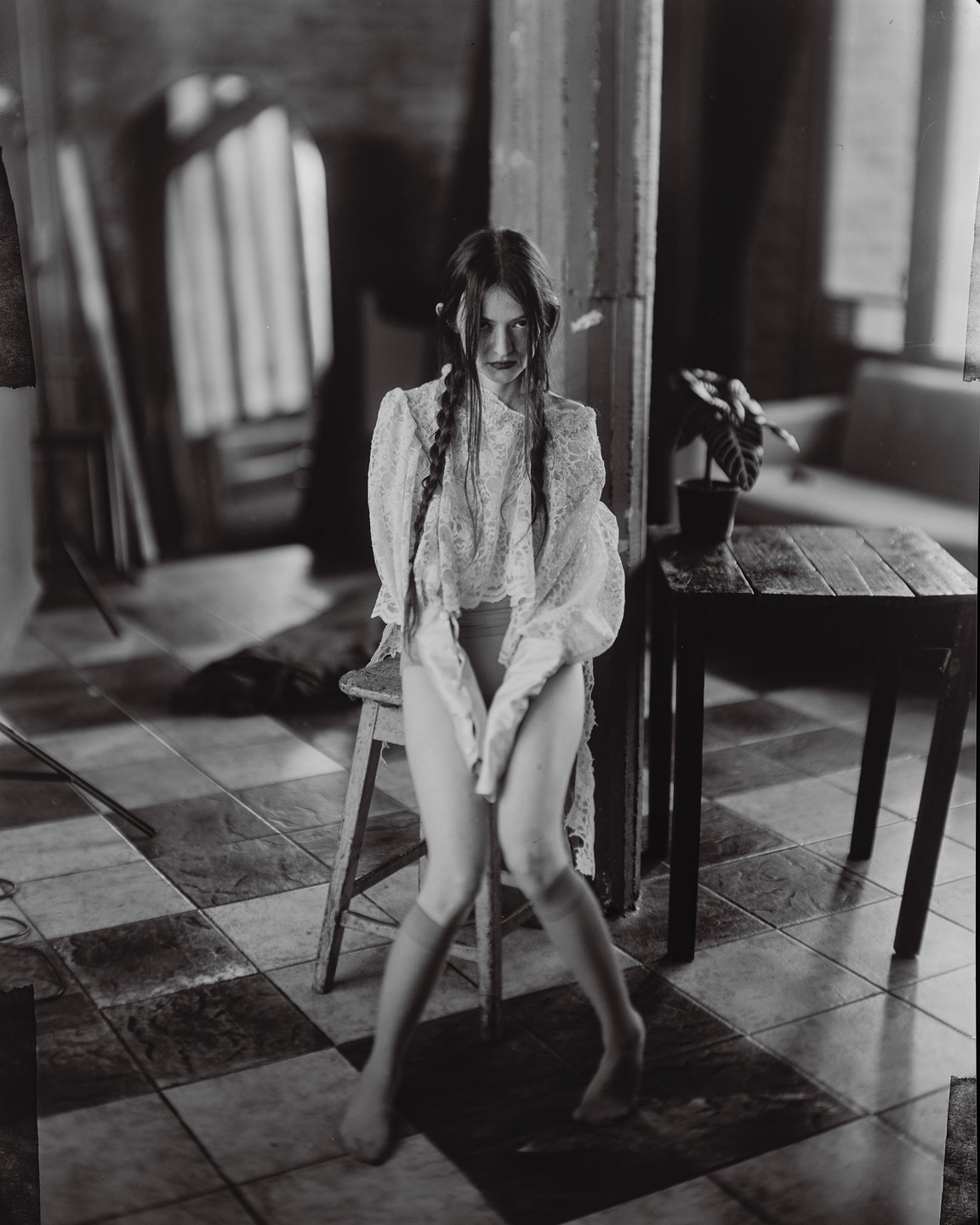A young woman with braided hair and a lace top sits on a stool in a room with a checkered floor.