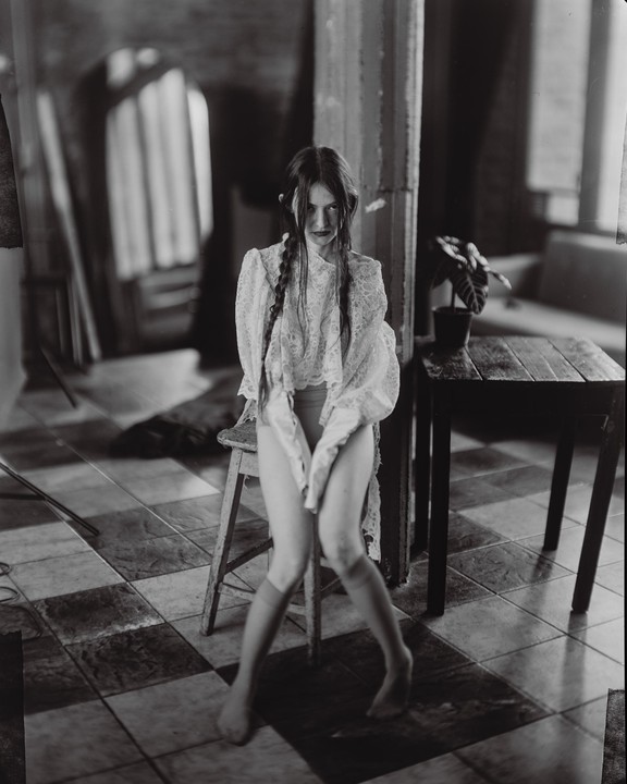 A young woman with braided hair and a lace top sits on a stool in a room with a checkered floor.