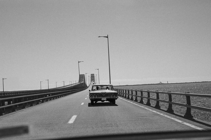 A car drives on a long bridge over water with a clear sky.