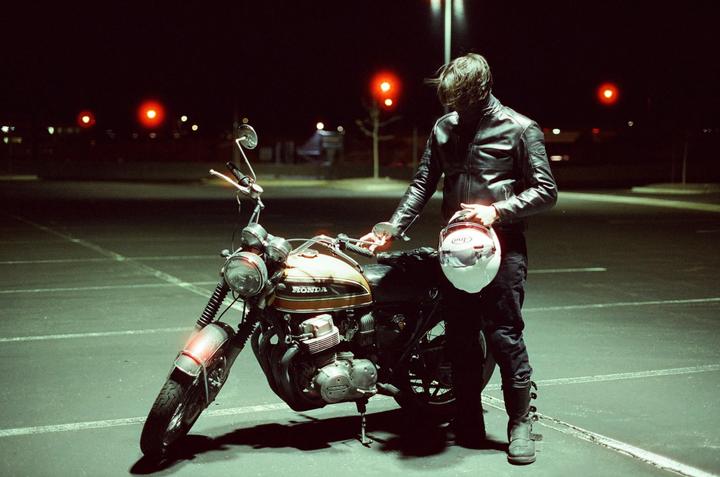 A man in a leather jacket stands next to a motorcycle at night, holding a helmet.