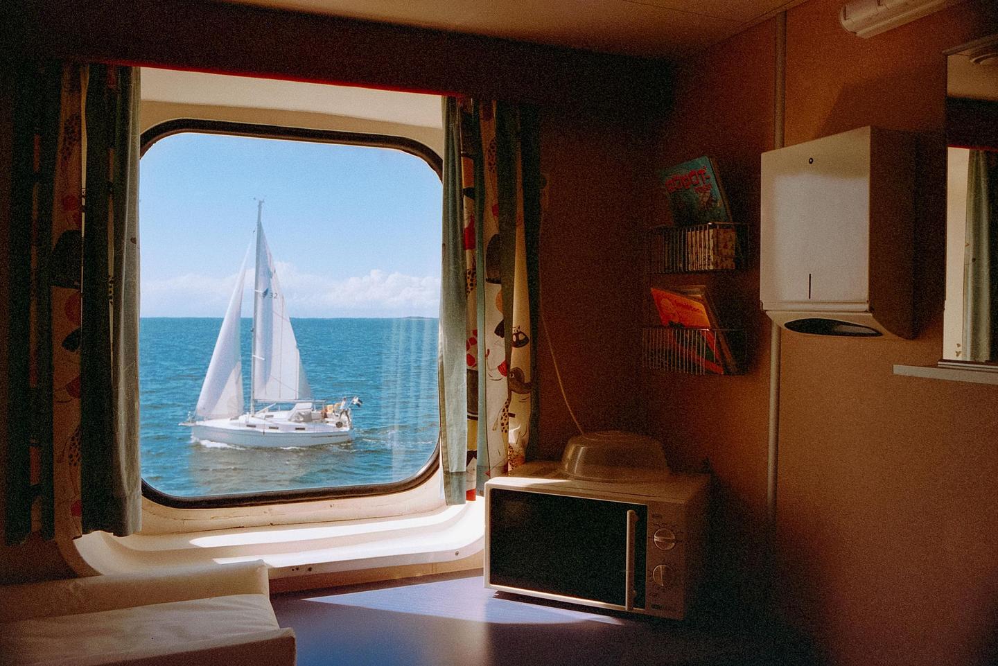 A sailboat is visible through a window from inside a boat cabin with a microwave and refrigerator.