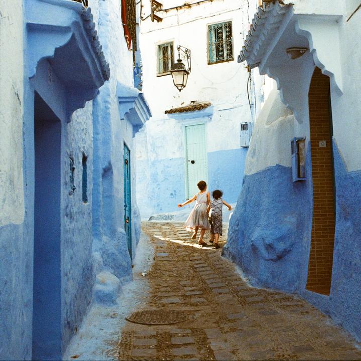 Two children walk down a narrow, blue-painted alleyway in a town.