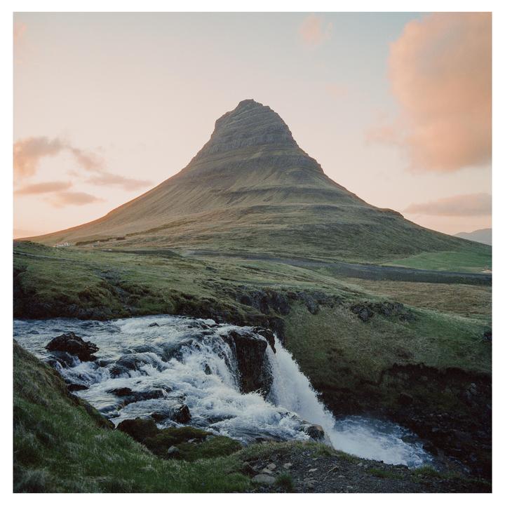 A waterfall flows over rocks in the foreground with a large mountain in the background under a sunset sky.
