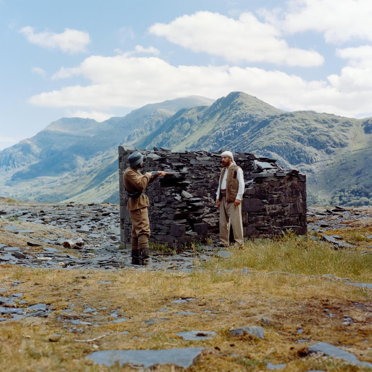Two men stand near a stone structure in a mountainous landscape.