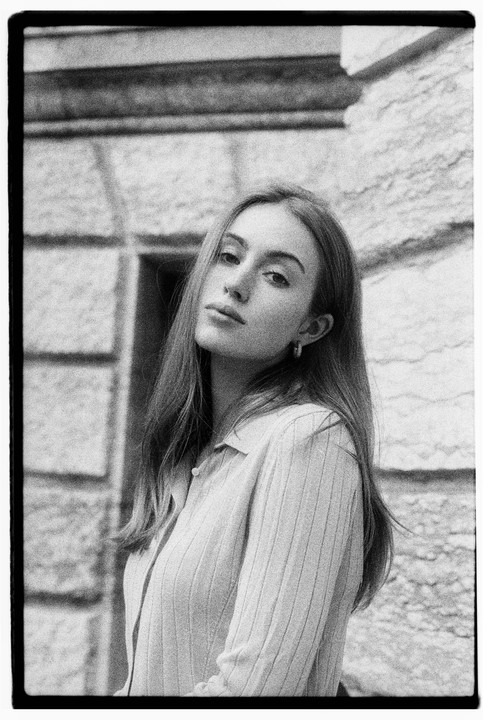 A woman with long hair and a collared shirt poses in front of a stone wall.