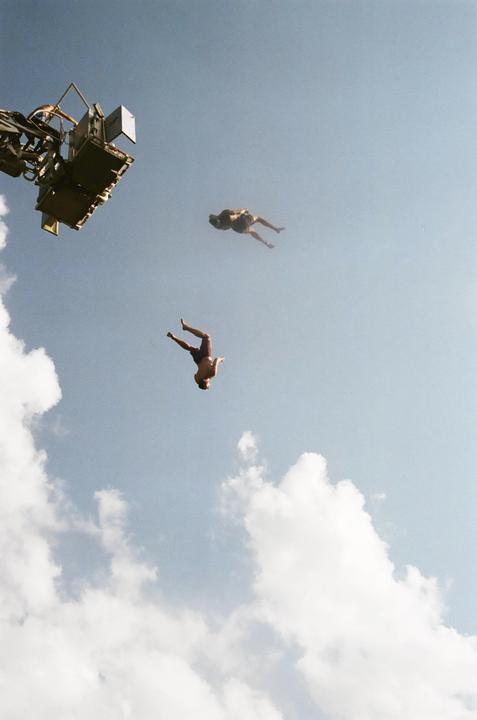 Two men are captured mid-air against a bright blue sky with scattered white clouds, one appearing to dive and the other floating.