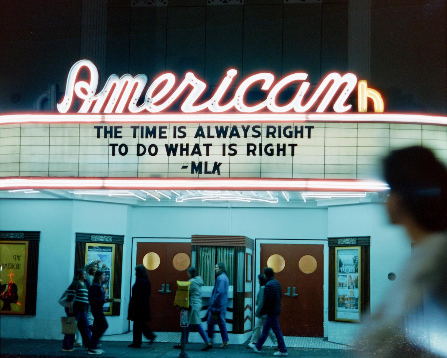 A group of people walk past a theater marquee that reads "American" and "The time is always right to do what is right - MLK" at night.