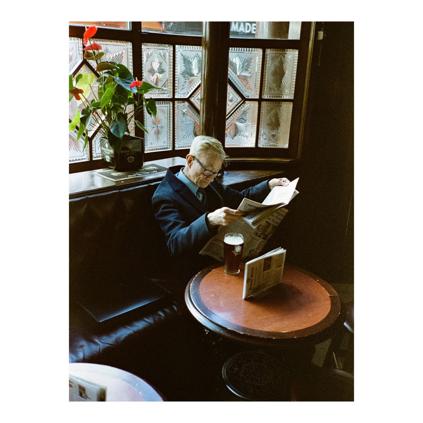 A man in a suit and glasses reads a newspaper at a table in a pub with a pint of beer.