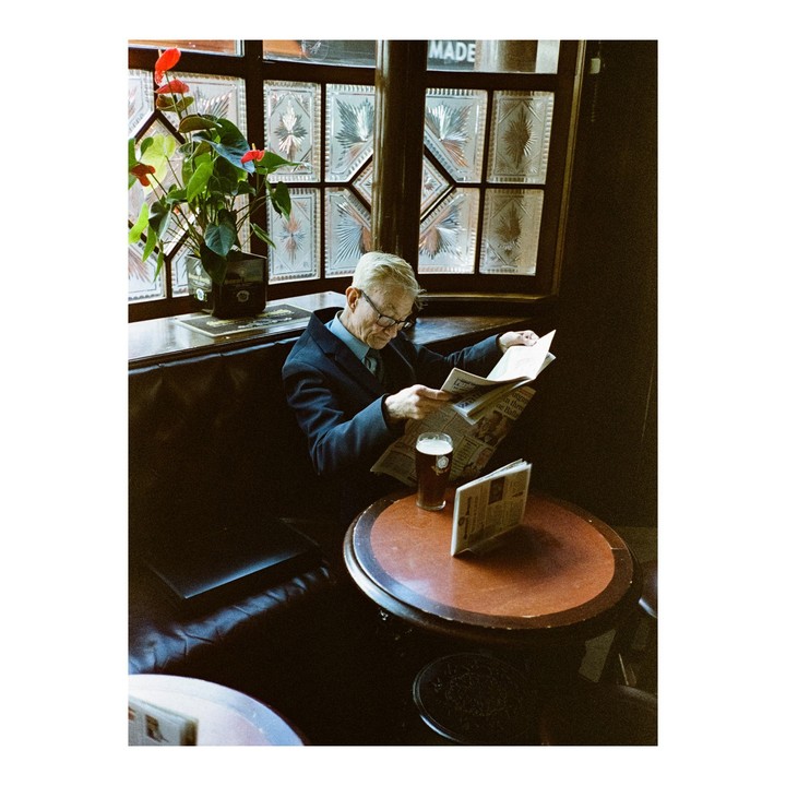 A man in a suit and glasses reads a newspaper at a table in a pub with a pint of beer.