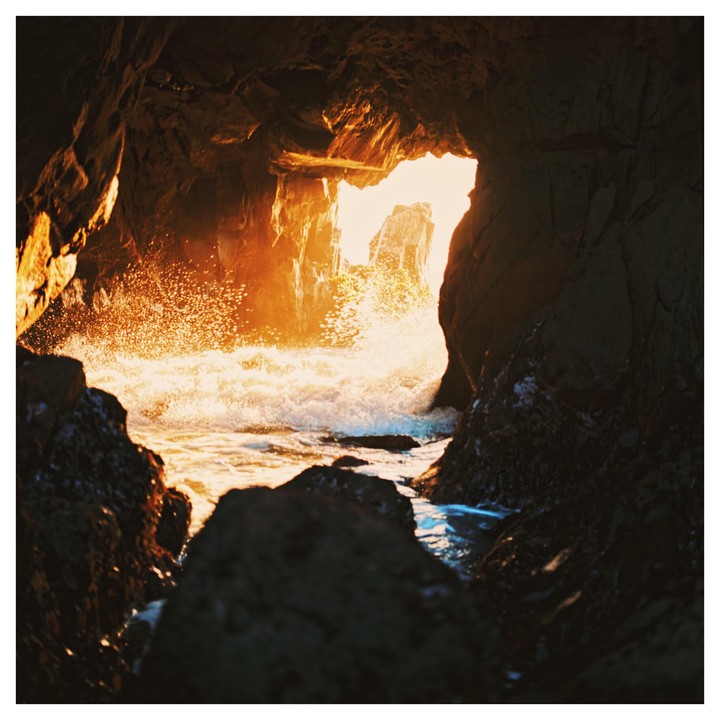 Waves crash against rocks inside a sea cave, with bright light visible through the cave opening.