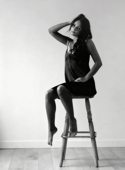 A woman in a dress sits on a stool with one arm raised to her head.