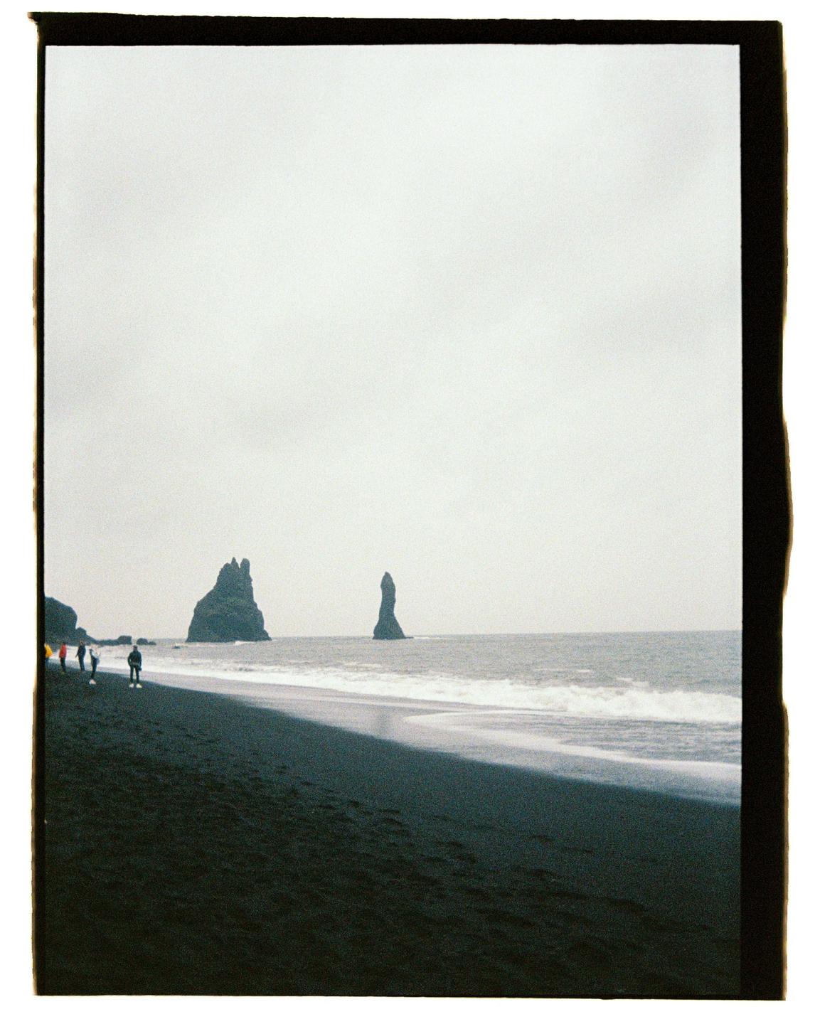 A group of people walk along a black sand beach with rock formations in the ocean under a cloudy sky.