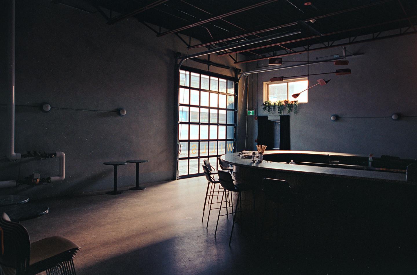A bar with stools and tables is illuminated by light coming through a large glass garage door and a window.