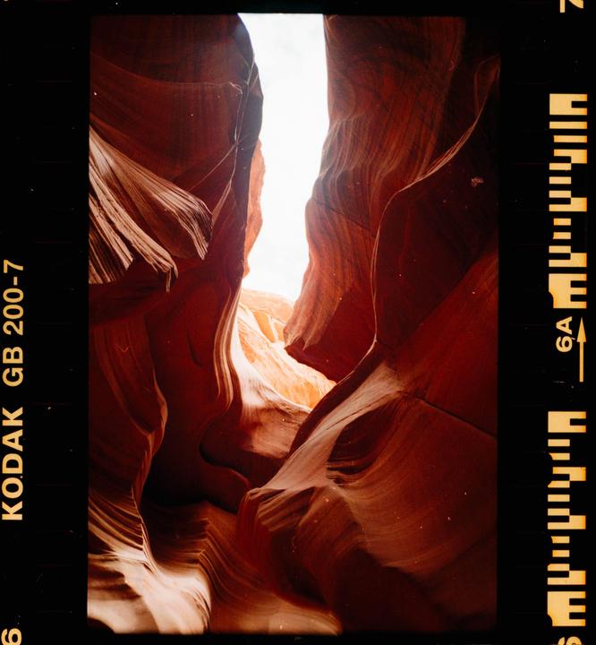 A narrow slot canyon with red and orange rock formations is illuminated by bright light from above.