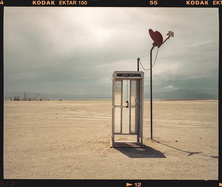 A telephone booth with a red heart antenna stands in a vast, barren landscape under a cloudy sky.