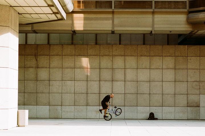 A man on a bicycle performs a trick against a tiled wall with sunlight and shadow.