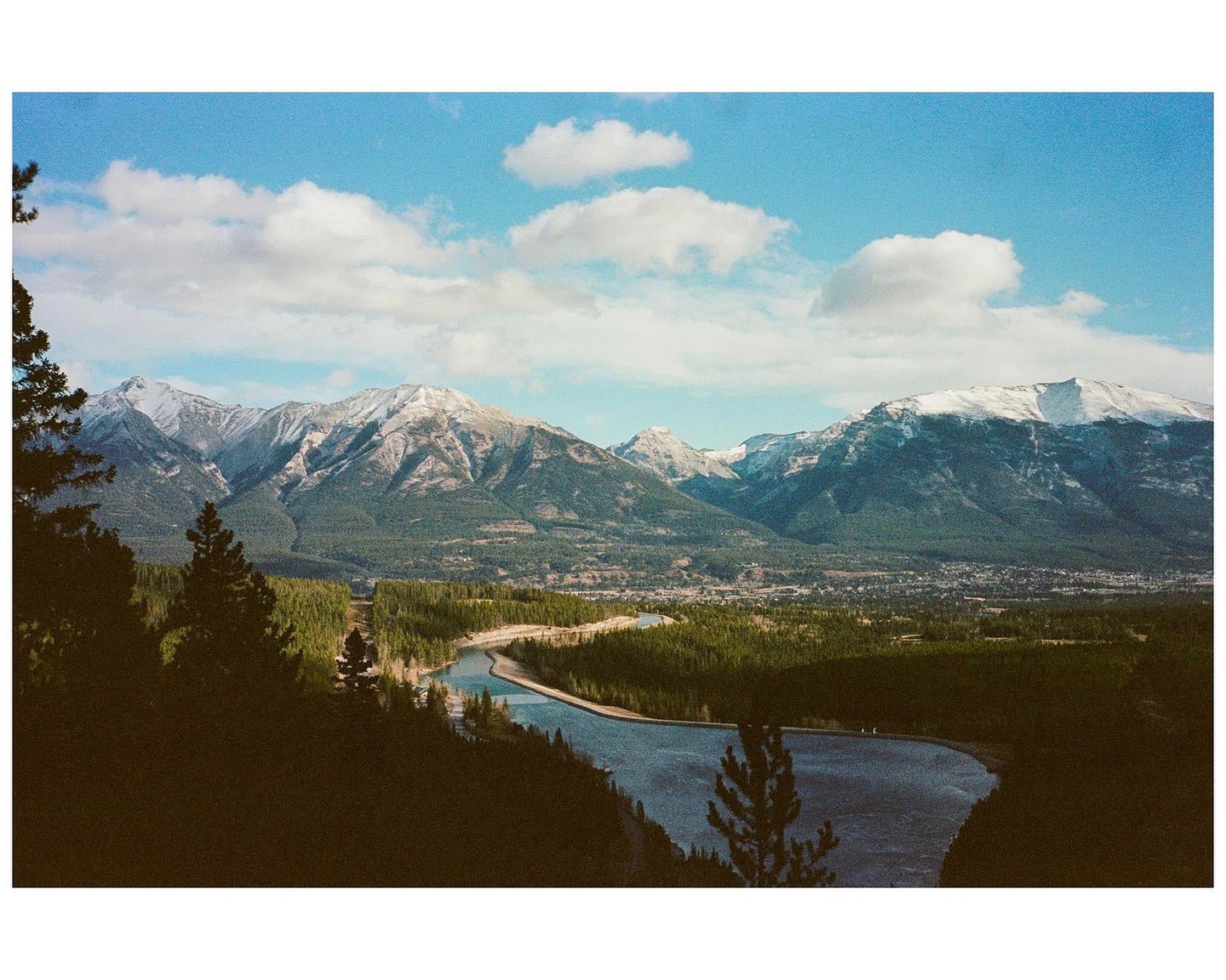 A river winds through a valley with a town and forest, surrounded by snow-capped mountains under a blue sky with clouds.