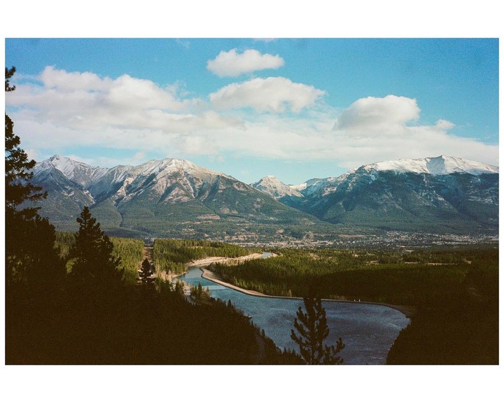 A river winds through a valley with a town and forest, surrounded by snow-capped mountains under a blue sky with clouds.