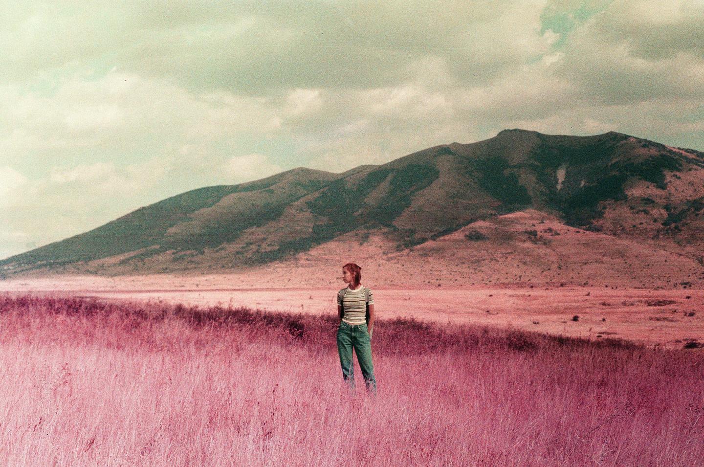 A woman stands in a field of purple grass with mountains in the background under a cloudy sky.