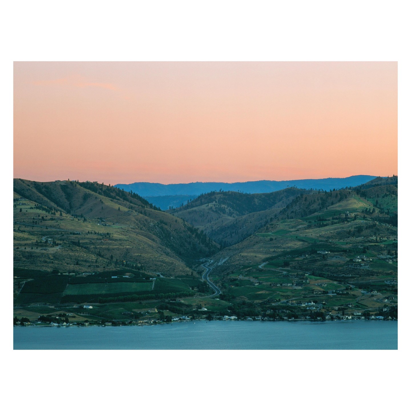 A winding road leads through a valley of green hills towards a body of water under a pink and blue sky.