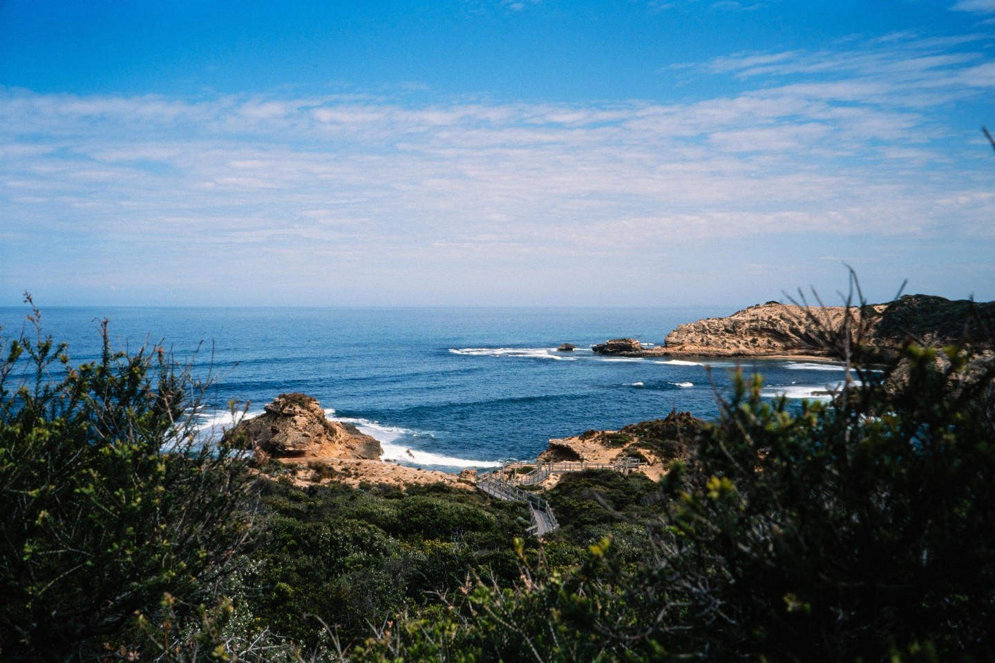 A coastal landscape features a blue ocean with waves crashing against rocky outcrops and a wooden boardwalk leading down to the shore, framed by green bushes in the foreground.