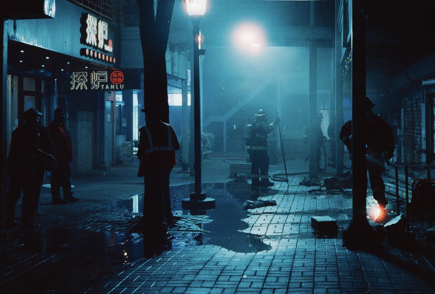 Firefighters and other people are gathered on a wet, brick-paved street at night, illuminated by streetlights and neon signs.