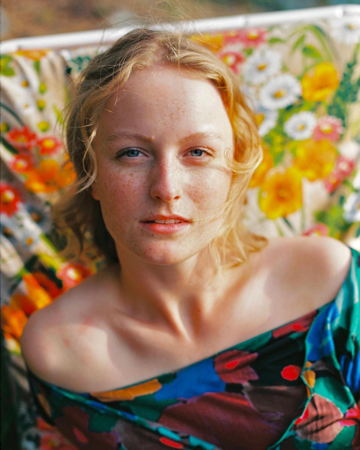 A woman with freckles and blonde hair looks at the viewer, wearing a floral top with a floral pattern in the background.