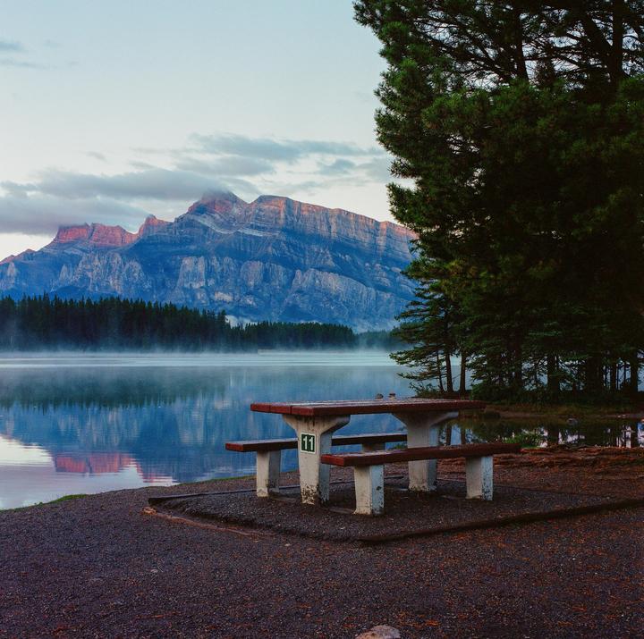 A picnic table sits on the shore of a lake with a forest and mountains in the background.