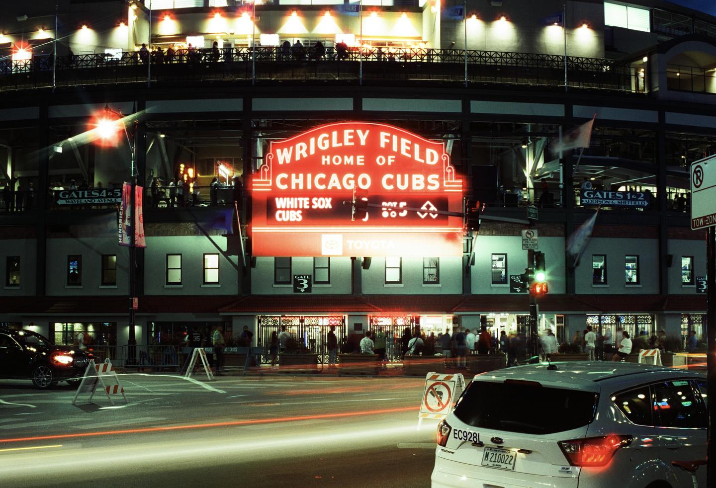 A large red sign for Wrigley Field and the Chicago Cubs is illuminated at night above a street with cars and people.