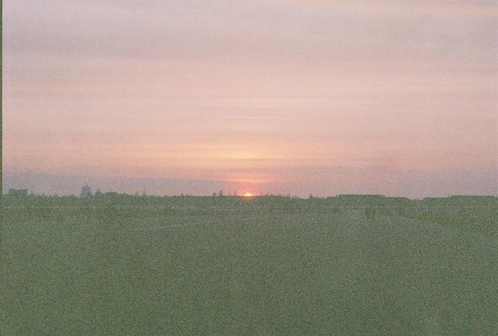 The sun sets over a field with a distant cityscape visible on the horizon.