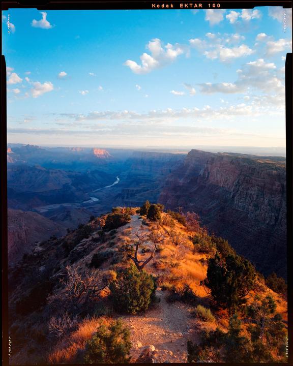A winding path leads along a ridge overlooking a vast canyon with a river at the bottom under a blue sky with clouds.