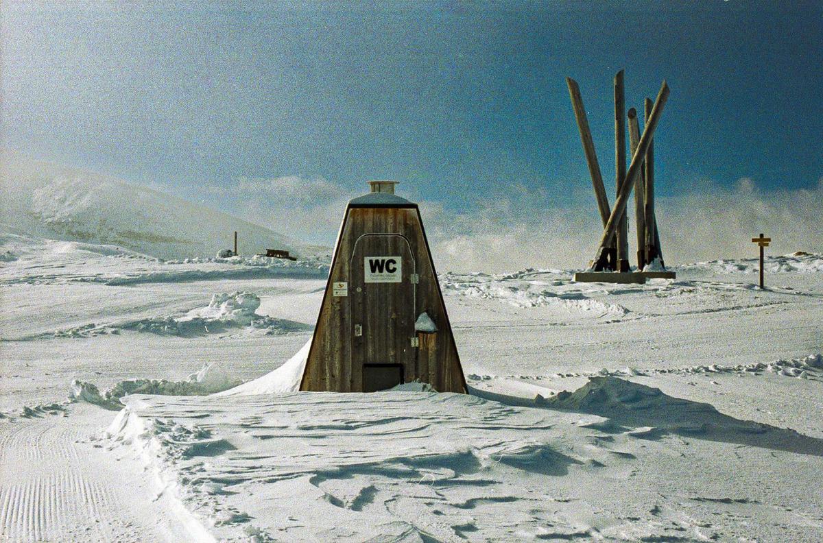A wooden outhouse stands in a snow-covered mountain landscape with a wooden sculpture and a signpost in the background.