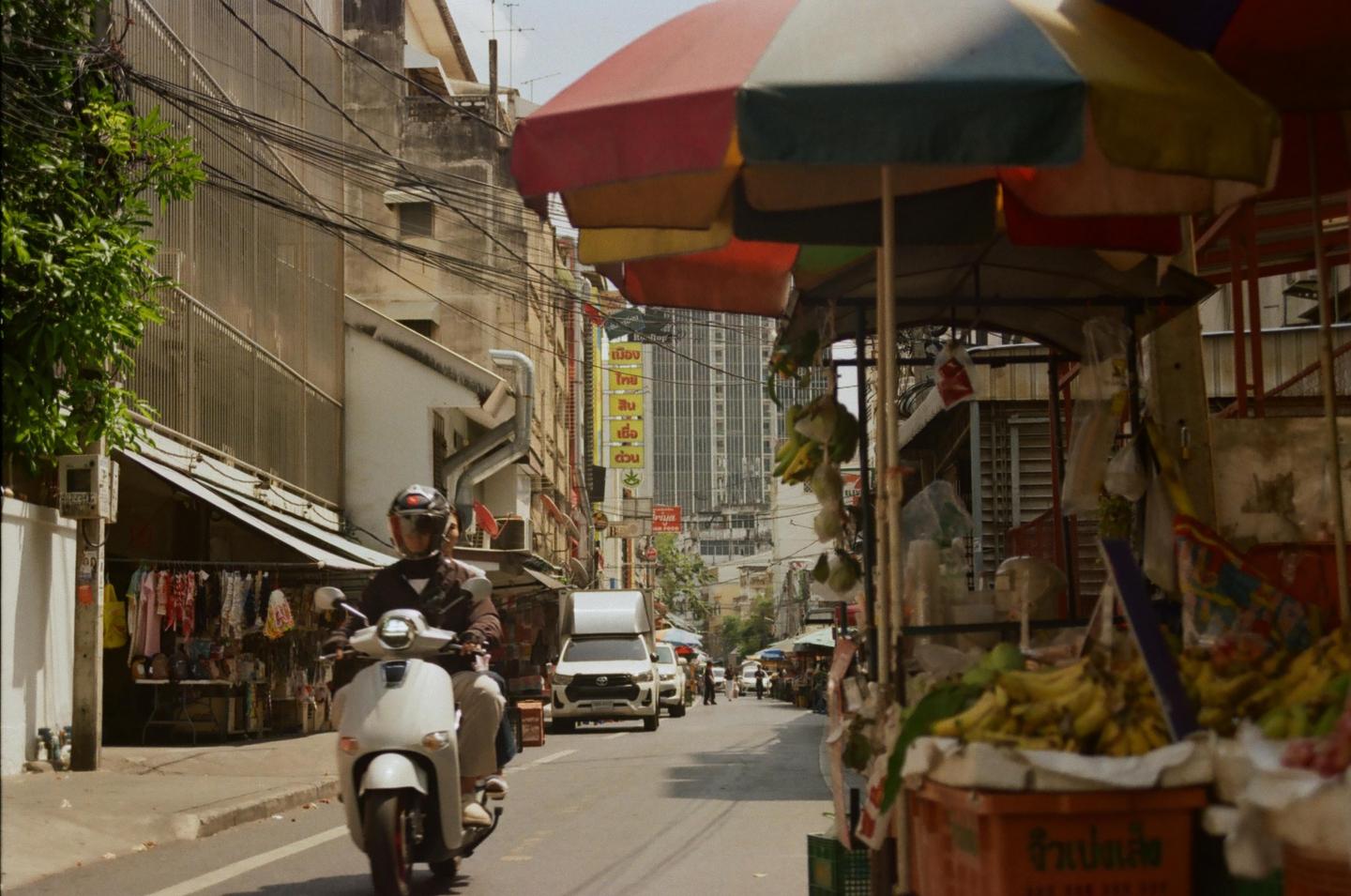 A person on a scooter drives down a busy street lined with market stalls and buildings in Bangkok.