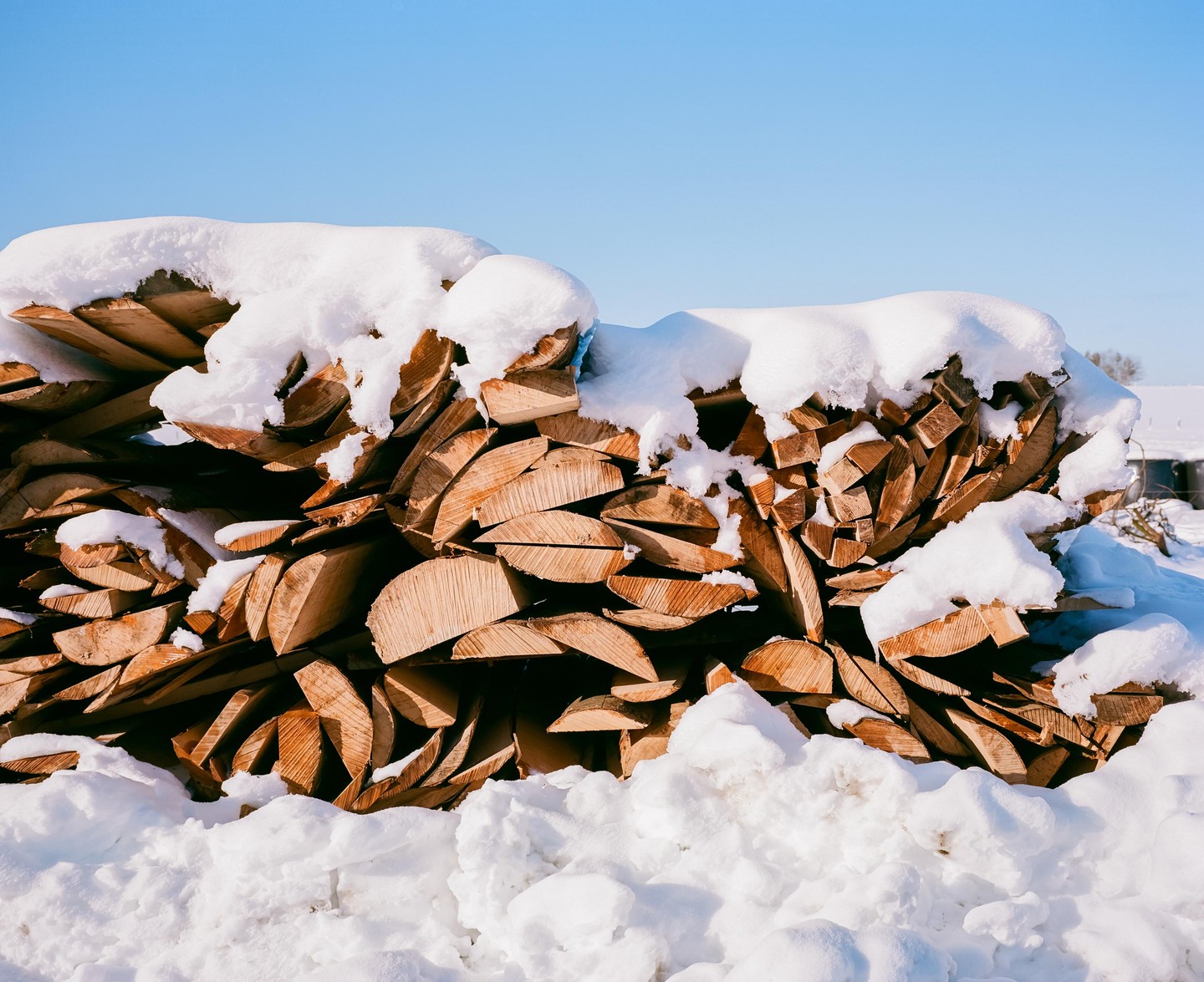 A large pile of split firewood is covered in snow under a clear blue sky.