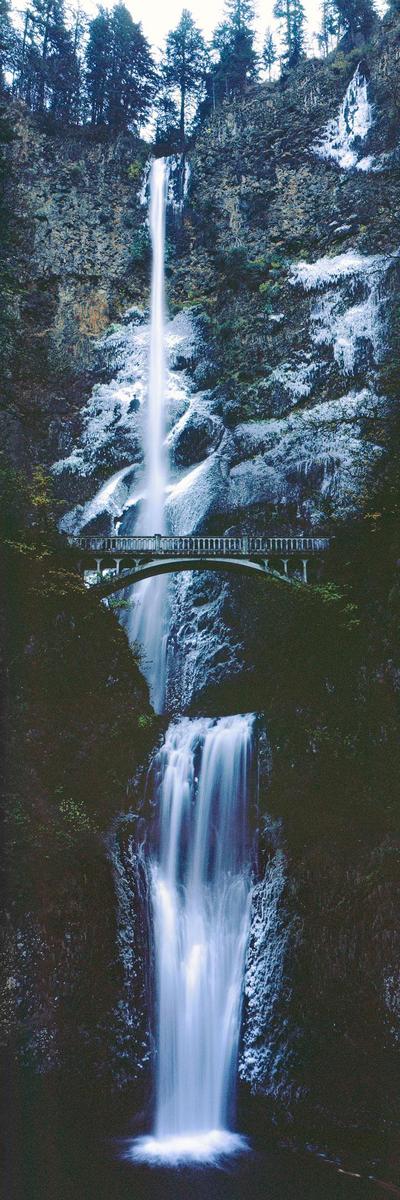 A tall waterfall cascades down a rocky cliff with a bridge crossing in front of it.