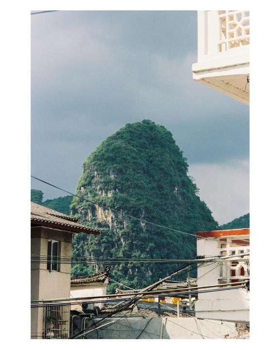 A lush green mountain rises behind buildings and a tangle of power lines under a cloudy sky.