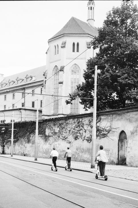 Three men ride electric scooters down a street next to a stone wall covered in ivy and a large building with a tall, pointed roof.