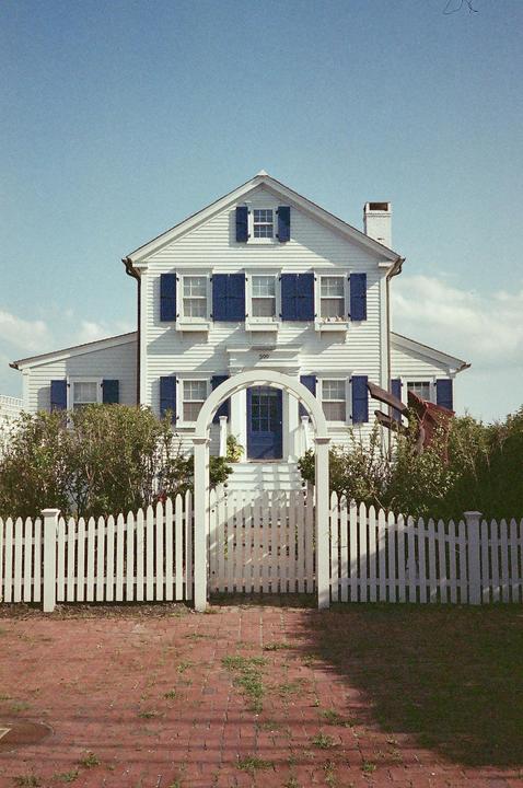 A white house with blue shutters and a blue door stands behind a white picket fence and brick path.