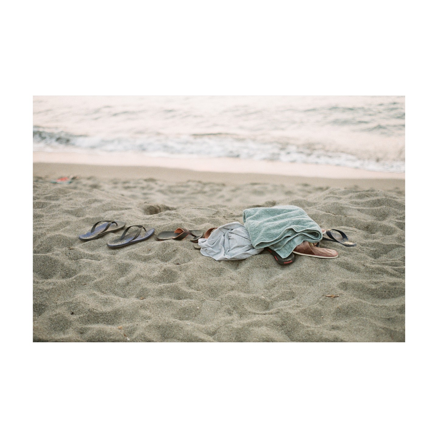 A towel, a shirt, and several pairs of sandals are left on a sandy beach near the ocean.