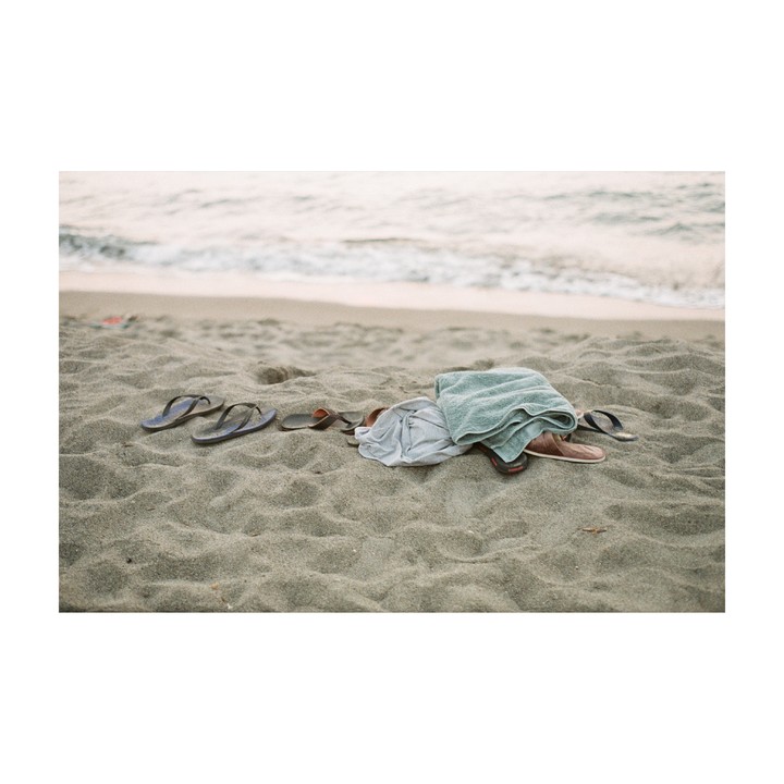 A towel, a shirt, and several pairs of sandals are left on a sandy beach near the ocean.
