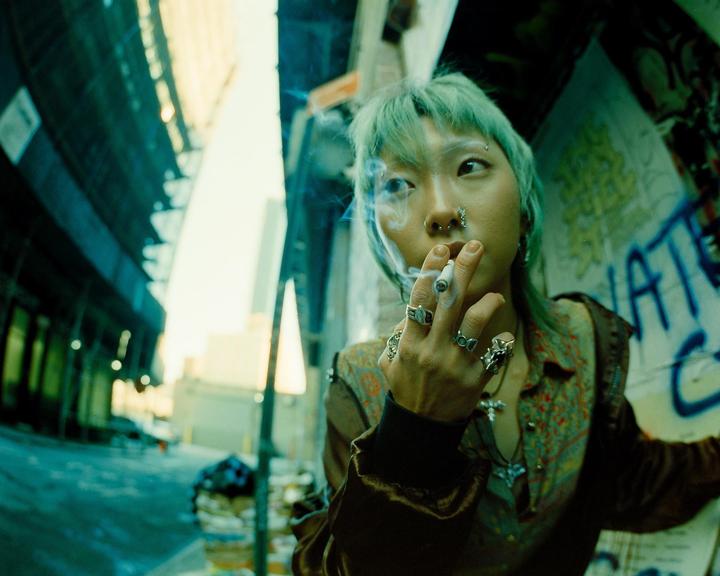 A person with green hair and multiple piercings smokes a cigarette in an alleyway with graffiti.