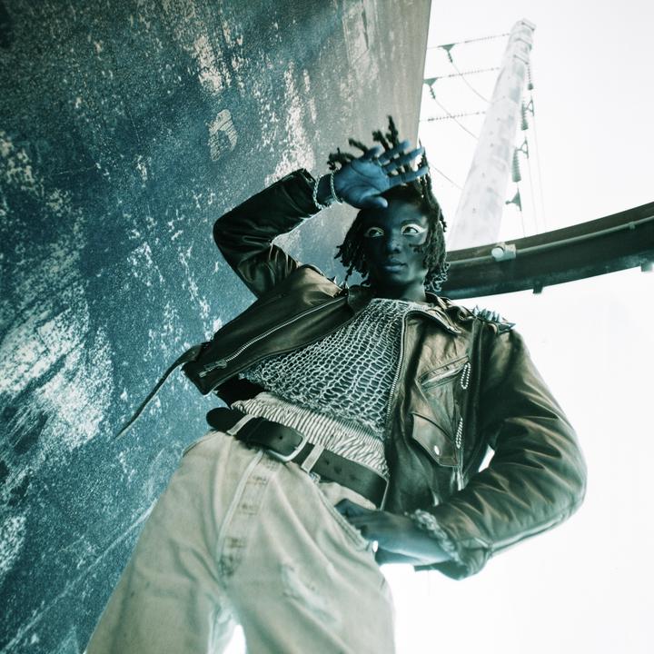 A person with dark skin and dreadlocks, wearing a leather jacket and light-colored pants, poses with one hand shielding their eyes.