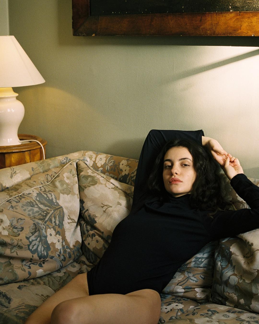 A woman in a black top is reclining on a floral sofa next to a lamp.