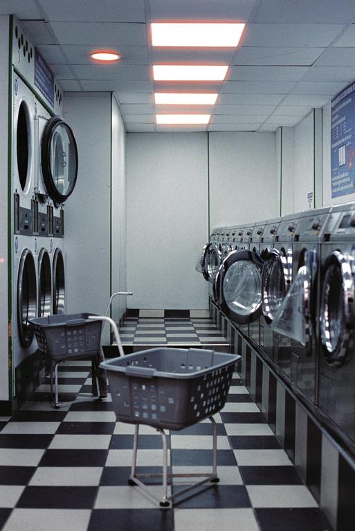 A long, narrow laundromat with a checkered floor is lined with washing machines and laundry baskets.