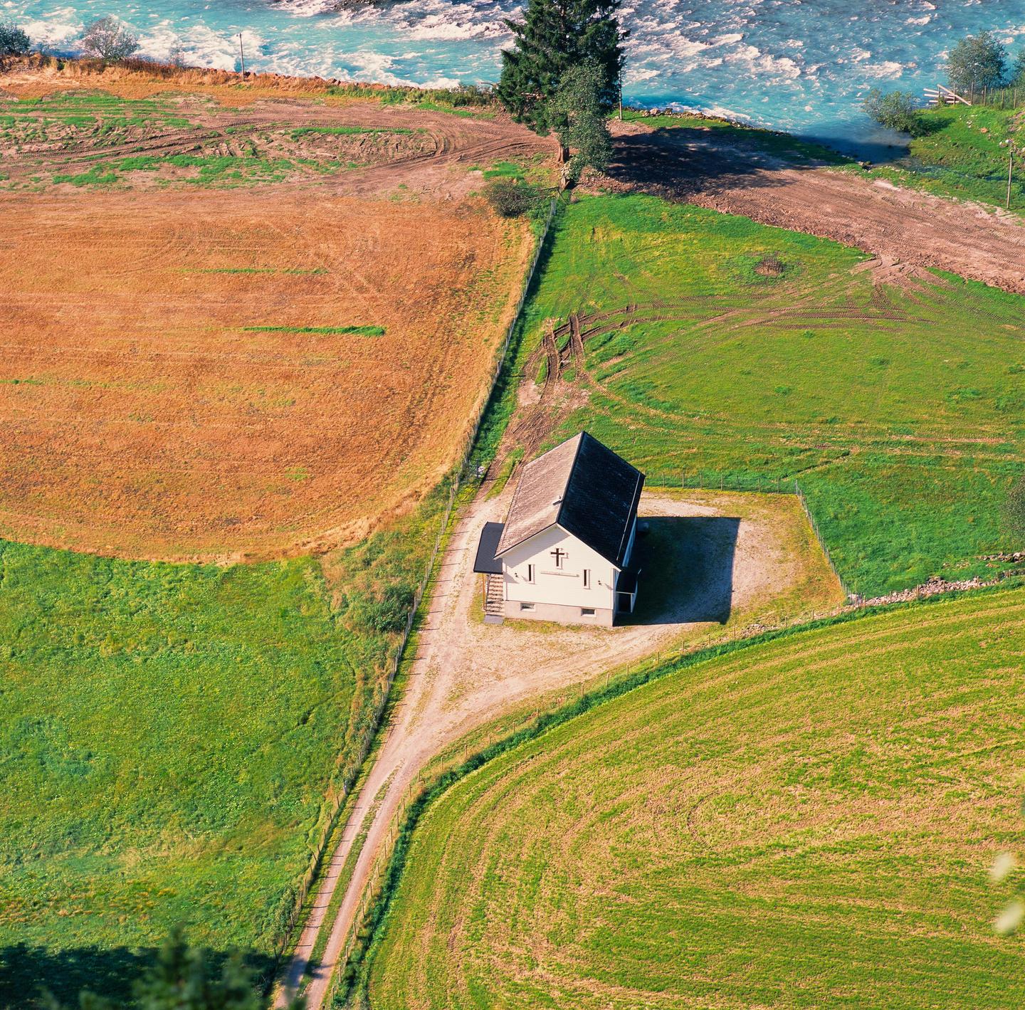 A small house with a cross on its side stands in a field next to a river.